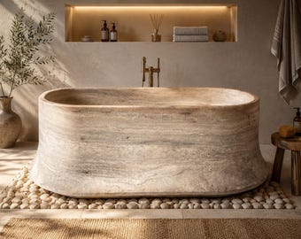Puede incluir: Una escena de baño lujosa con una gran bañera de piedra. La bañera se encuentra sobre un lecho de guijarros, con una alfombra tejida delante. Un pequeño taburete de madera contiene productos de baño. La habitación tiene una paleta de colores neutros y luz natural.