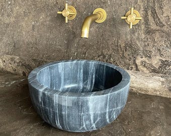 Puede incluir: Un lavabo de mármol gris oscuro con forma redonda. Cuenta con un grifo de latón y dos válvulas de cruz de latón. El lavabo está colocado contra una pared de mármol marrón.