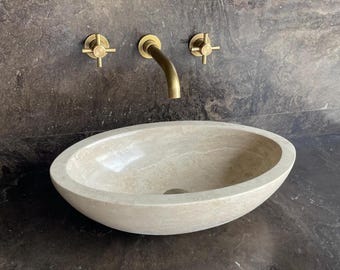 Puede incluir: Un lavabo ovalado de piedra beige con encimera y pared gris oscuro. El lavabo tiene un orificio de desagüe y se combina con grifería de latón, incluyendo un grifo curvo y dos manijas en forma de cruz. Diseño moderno y minimalista.