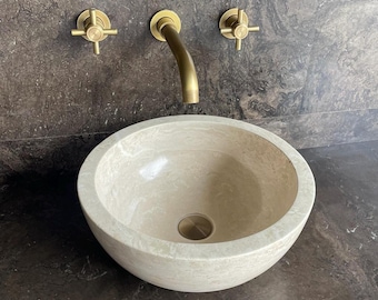 Puede incluir: Un lavabo de piedra beige de forma redonda con un acabado liso y pulido y un desagüe central. Sobre el lavabo, un grifo de latón montado en la pared presenta un caño curvo flanqueado por dos mandos en cruz sobre una pared de piedra oscura.