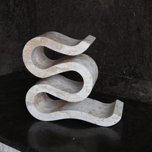 Puede incluir: Una escultura abstracta de color claro, con un dise&ntilde;o sinuoso y fluido. La escultura est&aacute; hecha de un material similar a la piedra y se encuentra sobre una superficie oscura y reflectante. La pieza tiene una est&eacute;tica moderna y art&iacute;stica.