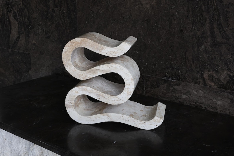Puede incluir: Una escultura abstracta de color claro, con un dise&ntilde;o sinuoso y fluido. La escultura est&aacute; hecha de un material similar a la piedra y se encuentra sobre una superficie oscura y reflectante. La pieza tiene una est&eacute;tica moderna y art&iacute;stica.