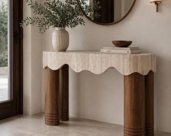 Puede incluir: Una consola decorativa con borde festoneado y tres patas cilíndricas de madera. La mesa tiene un jarrón con vegetación, un cuenco de madera y libros. La mesa es de color neutro y las patas son de color marrón oscuro. La mesa mide unos 100 cm de altura.