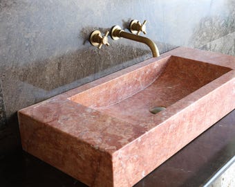 Puede incluir: Un lavabo rectangular de mármol rojo-anaranjado con una cubeta interior curvada. El lavabo tiene una encimera oscura y se combina con un grifo dorado y manijas en forma de cruz. El fondo presenta una pared gris oscuro texturizada.