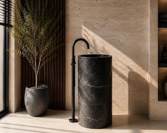 Puede incluir: Un lavabo de pedestal de mármol negro con un grifo negro a juego. El lavabo cilíndrico se apoya sobre un suelo de color claro. A la izquierda del lavabo hay una planta en maceta y detrás un panel de madera. La pared es de color beige claro.