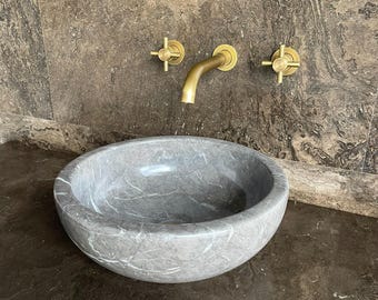 Puede incluir: Un lavabo de mármol gris con forma redondeada se encuentra sobre una encimera marrón oscuro. Encima del lavabo, un grifo dorado y dos manijas en forma de cruz están montados en una pared de mármol marrón. El lavabo tiene vetas blancas.
