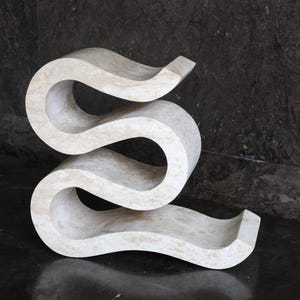 Puede incluir: Una escultura abstracta de color crema con un dise&ntilde;o sinuoso y fluido. La escultura tiene una superficie lisa y pulida y est&aacute; hecha de un material s&oacute;lido similar a la piedra. La pieza se exhibe sobre una superficie oscura y reflectante.