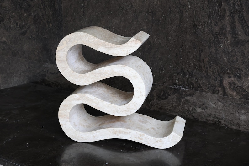 Puede incluir: Una escultura abstracta de color crema con un dise&ntilde;o sinuoso y fluido. La escultura tiene una superficie lisa y pulida y est&aacute; hecha de un material s&oacute;lido similar a la piedra. La pieza se exhibe sobre una superficie oscura y reflectante.