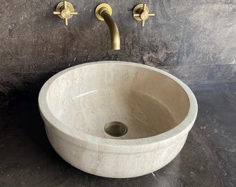Puede incluir: Un lavabo redondo beige con un grifo de latón cepillado y manijas a juego. El lavabo está colocado contra una pared oscura y texturizada. El grifo tiene un caño curvo. Las manijas tienen forma de cruz.