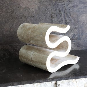 Puede incluir: Escultura abstracta en forma de S, hecha de un material beige claro con un acabado liso. Se encuentra sobre una superficie oscura y reflectante, contra un fondo gris texturizado. Una obra de arte moderna.