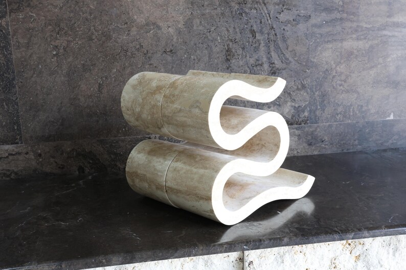 Puede incluir: Escultura abstracta en forma de S, hecha de un material beige claro con un acabado liso. Se encuentra sobre una superficie oscura y reflectante, contra un fondo gris texturizado. Una obra de arte moderna.