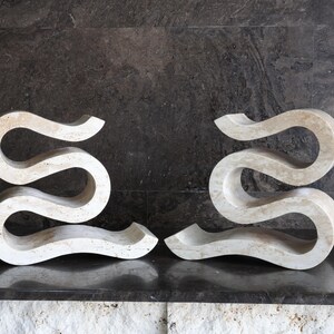 Puede incluir: Dos esculturas abstractas en color beige claro. Cada escultura tiene una forma serpentina fluida, con una base plana y una parte superior curva. Las esculturas est&aacute;n hechas de un material similar a la piedra y se exhiben sobre una superficie oscura.
