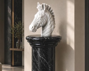 Puede incluir: Una escultura de cabeza de cebra blanca con rayas talladas en relieve y una crin texturizada. La escultura está colocada sobre un pedestal cilíndrico de mármol negro con vetas blancas, que presenta un acabado liso y pulido y un borde superior redondeado.