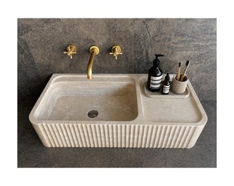 Puede incluir: Un lavabo rectangular beige con un diseño frontal acanalado. El lavabo tiene un estante incorporado con un dispensador de jabón, una botella pequeña y un soporte para cepillos de dientes. Los grifos y accesorios de pared dorados están montados encima del lavabo.