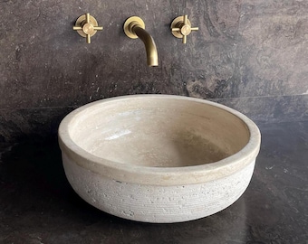 Puede incluir: Un lavabo redondo de piedra clara con un exterior texturizado. El lavabo está colocado contra una pared y encimera oscuras y moteadas, con grifos de latón encima. El grifo tiene un caño curvo y dos manijas en forma de cruz.