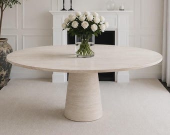 Puede incluir: Una mesa de comedor redonda de travertino en color beige claro. La mesa tiene una base de pedestal y una superficie circular lisa. Un jarrón con rosas blancas está sobre la mesa. El texto "ROUND TRAVERTINE DINING TABLE" está en la parte inferior de la imagen.