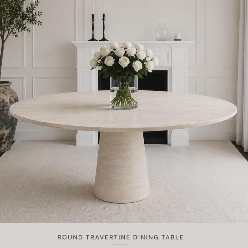 May include: A circular travertine dining table in a light beige hue. The table features a pedestal base and a smooth, round surface. A vase of white roses is placed on the table. The text "ROUND TRAVERTINE DINING TABLE" is at the bottom of the image.