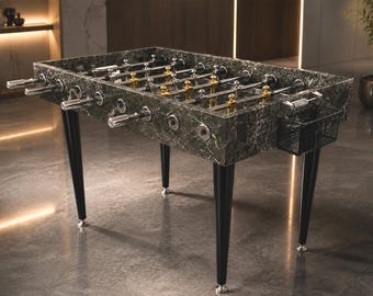 Puede incluir: Una mesa de futbolín con un mueble de patrón de mármol verde oscuro, cuatro patas negras cónicas con pies plateados y barras cromadas con figuras de jugadores doradas y plateadas. Una cesta de malla negra está fijada al lateral del mueble.