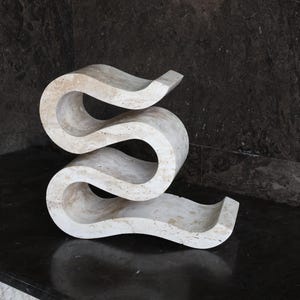 Puede incluir: Una escultura abstracta de color crema con un dise&ntilde;o curvo y fluido. La obra de arte de piedra tiene una textura suave y se asienta sobre una superficie oscura y reflectante. La forma de la escultura se asemeja a una 'S' alargada.