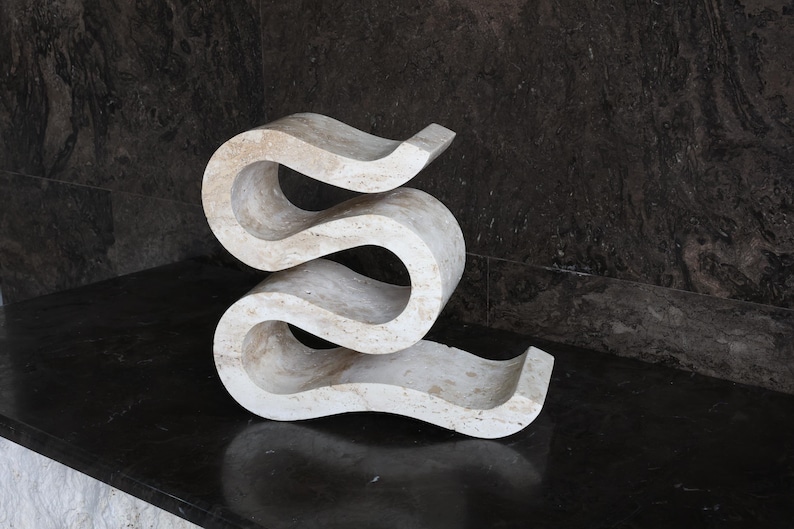 Puede incluir: Una escultura abstracta de color crema con un dise&ntilde;o curvo y fluido. La obra de arte de piedra tiene una textura suave y se asienta sobre una superficie oscura y reflectante. La forma de la escultura se asemeja a una 'S' alargada.