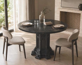 Puede incluir: Una mesa de comedor redonda de mármol negro con una base de pedestal arquitectónica detallada, acompañada de dos sillas con marcos de madera oscura y asientos tapizados en color beige claro. La mesa tiene platos y un jarrón.