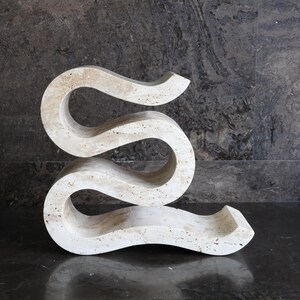 Puede incluir: Una escultura abstracta de color crema con un dise&ntilde;o fluido y serpentino. La talla de piedra tiene un acabado liso y pulido y se exhibe sobre un fondo oscuro y texturizado. La forma de la escultura recuerda a una "S" alargada.