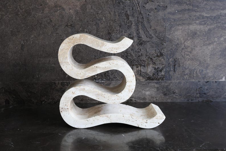 Puede incluir: Una escultura abstracta de color crema con un dise&ntilde;o fluido y serpentino. La talla de piedra tiene un acabado liso y pulido y se exhibe sobre un fondo oscuro y texturizado. La forma de la escultura recuerda a una "S" alargada.