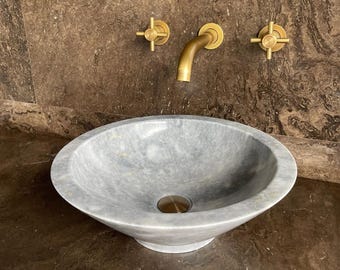Puede incluir: Un lavabo ovalado de mármol gris con grifería de latón. El lavabo tiene un desagüe de latón y está sobre una superficie marrón oscuro. El grifo y los mandos son de color dorado.
