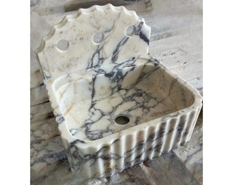 Puede incluir: Un lavabo de mármol blanco con vetas grises y doradas. El cuenco presenta un borde superior festoneado, un diseño exterior acanalado y un respaldo alto con tres orificios circulares para la grifería y un orificio central para el desagüe.