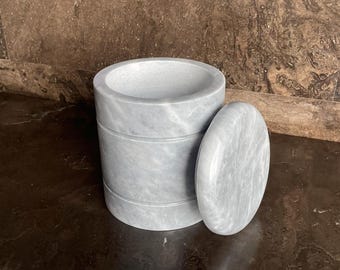 Puede incluir: Un juego de mortero y maja de mármol gris. El mortero es cilíndrico con ranuras horizontales, y la maja es una piedra plana y redonda. El juego está hecho de mármol gris claro con vetas grises más oscuras.