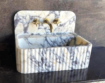 Puede incluir: Un lavabo de mármol con un patrón veteado blanco y gris. El lavabo tiene una cubeta rectangular y un protector contra salpicaduras curvado. Cuenta con grifos de latón, incluyendo un grifo y dos manijas. El exterior del lavabo tiene un diseño acanalado.