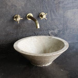 May include: A stone washbasin with brass taps and a spout, set against a dark grey wall. The basin is light beige and rounded. The taps and spout are a warm gold colour.