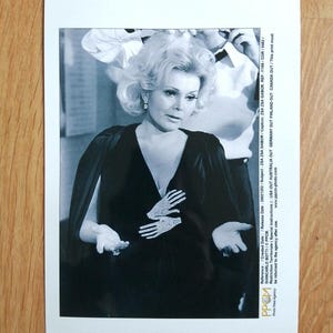May include: A black and white photograph of Zsa Zsa Gabor wearing a dark V-neck dress with a prominent, light-colored, hand-shaped embellishment on the chest. The image includes printed text along the right margin detailing agency information.