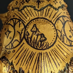 Puede incluir: Un cráneo pintado de oro con un diseño de tinta negra que presenta tres lunas, estrellas y hongos. El diseño recuerda un tema celestial o místico.