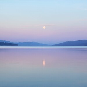 Op de afbeelding: Een volle maan hangt aan een lichtblauwe hemel boven een stil meer en reflecteert in het water. De bergen in de verte zijn gesilhouetteerd tegen de roze en paarse lucht.