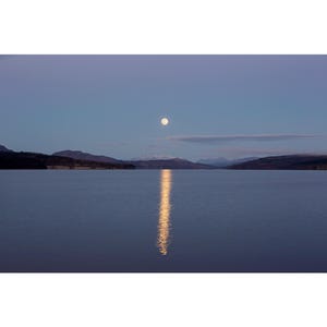 Op de afbeelding: Een volle maan hangt aan een lichtblauwe hemel boven een stil meer, en reflecteert een heldere lichtstraal op het water. In de verte zijn bergen te zien.