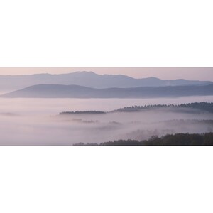 May include: A panoramic view of a misty landscape with rolling hills and a distant mountain range. The fog creates a soft, ethereal atmosphere, with the hills and mountains appearing as silhouettes against the pale sky.