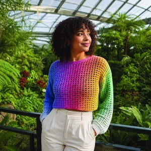 Könnte beinhalten: Ein offener Strickpullover in Regenbogenfarben, der von Blau über Lila zu Gelb und Grün verläuft. Das Model trägt den Pullover mit einer cremefarbenen Hose. Der Hintergrund ist ein üppiger grüner Garten.