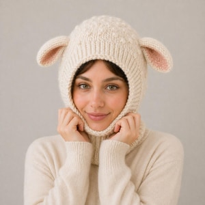 Op de afbeelding: Een crèmekleurige gebreide muts met aangehechte lammerenoren en een nekwarmer. De binnenkant van de oren van de muts is zachtroze. De persoon op de foto draagt een crèmekleurige trui en de achtergrond is effen lichtgrijs.