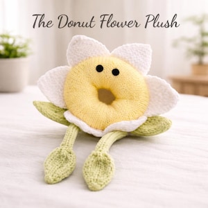 May include: A plush toy shaped like a flower with a donut center. The donut is yellow with a hole in the middle and black dot eyes. The flower petals are white, and the leaves and stems are green. The text "The Donut Flower Plush" is at the top.
