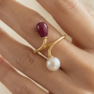Könnte beinhalten: Ein offener Ring mit einem mattgoldenen Band. Ein Ende des Bandes ist mit einem glatten, tropfenförmigen, tiefroten Edelstein besetzt, während das andere Ende eine runde, glänzende weiße Perle von ähnlicher Größe aufweist.