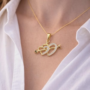 May include: A gold-toned necklace featuring a pendant with two interlocking hearts pierced by an arrow. The hearts are embellished with small, clear, sparkling stones set along the surface. The pendant hangs from a delicate gold-toned chain.