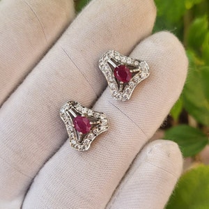 Puede incluir: Un par de pendientes de plata con un diseño triangular. Cada pendiente tiene una piedra preciosa roja rodeada de piedras blancas más pequeñas.
