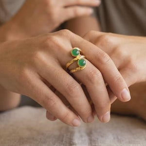 Può includere: Un anello aperto color oro con due pietre cabochon rotonde e lisce di colore verde incastonate alle estremità. La fascia in metallo presenta una finitura martellata e testurizzata che avvolge il dito, creando un design aperto distintivo.