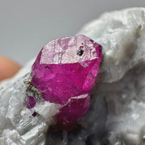 May include: A vibrant, deep pink, translucent crystal formation embedded within a rough, pale gray, crystalline rock matrix. The primary crystal features distinct geometric facets and a small, dark, circular inclusion on its top surface.