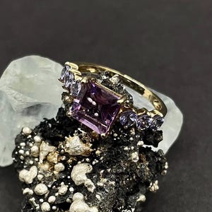 May include: A gold ring with a large, square-cut purple gemstone and smaller, round purple stones. The ring is set on a dark, textured surface. The background is a light colour.