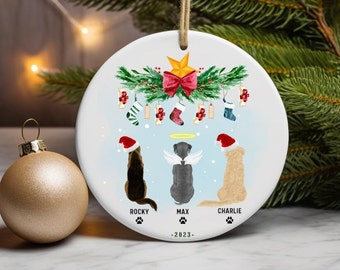 May include: White ceramic Christmas ornament with a green wreath and red bow at the top. Three dogs are depicted wearing Santa hats and standing under the wreath. The dogs are labeled "Rocky", "Max", and "Charlie". The ornament also includes the text "2023".