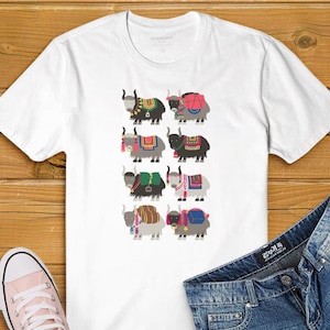 Op de afbeelding: Wit T-shirt met een kleurrijke illustratie van acht yaks die verschillende gepatroneerde zadeltassen dragen. De yaks kijken allemaal naar voren en hebben zwarte hoorns.