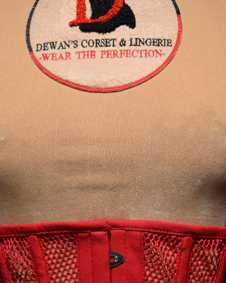 May include: A red corset with a black mesh panel and a silver metal hook and eye closure. The corset is worn over a beige shirt with a red and black embroidered patch that reads "Dewan's Corset & Lingerie -Wear the Perfection-"