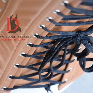May include: A brown leather corset with black laces and silver eyelets. The corset is laced up the back and has a logo that says "Dewan's Corset & Lingerie - Wear the Perfection - "